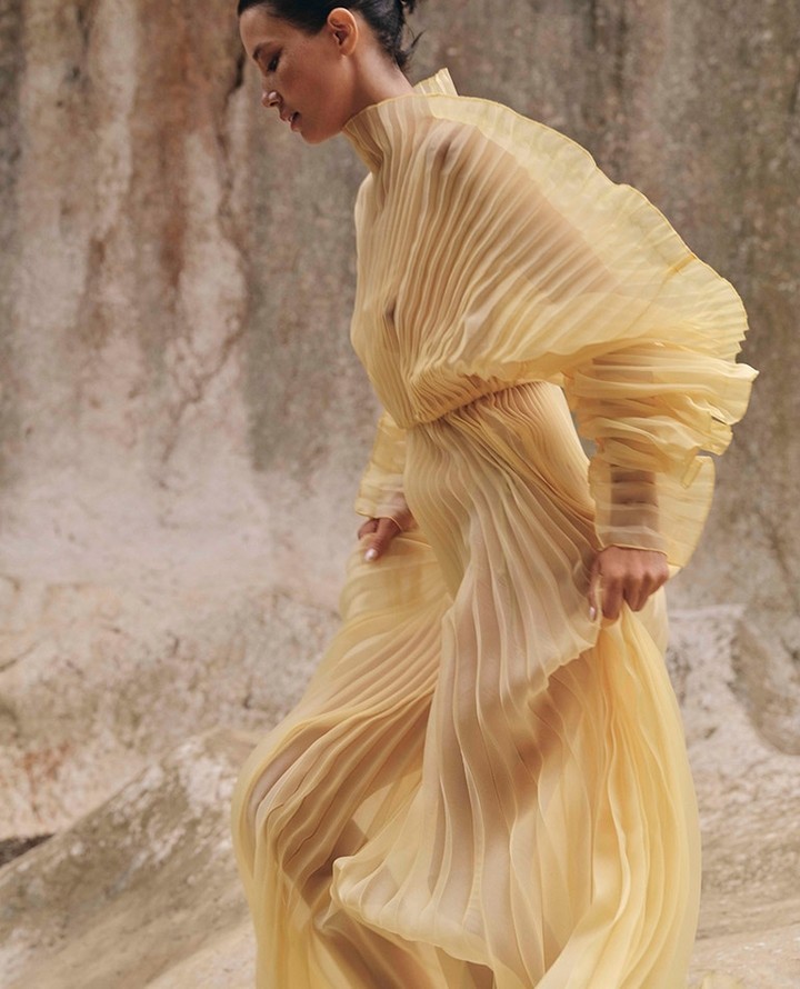 Woman in flowing yellow gown with pleated details, standing against a light tan textured surface, fashionable and elegant, walking.