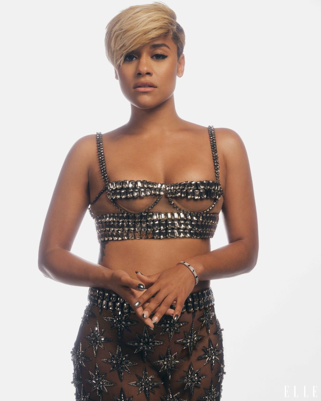 A woman with short blond hair poses in a studded bra and star-patterned skirt against a white backdrop, gazing directly at the camera.