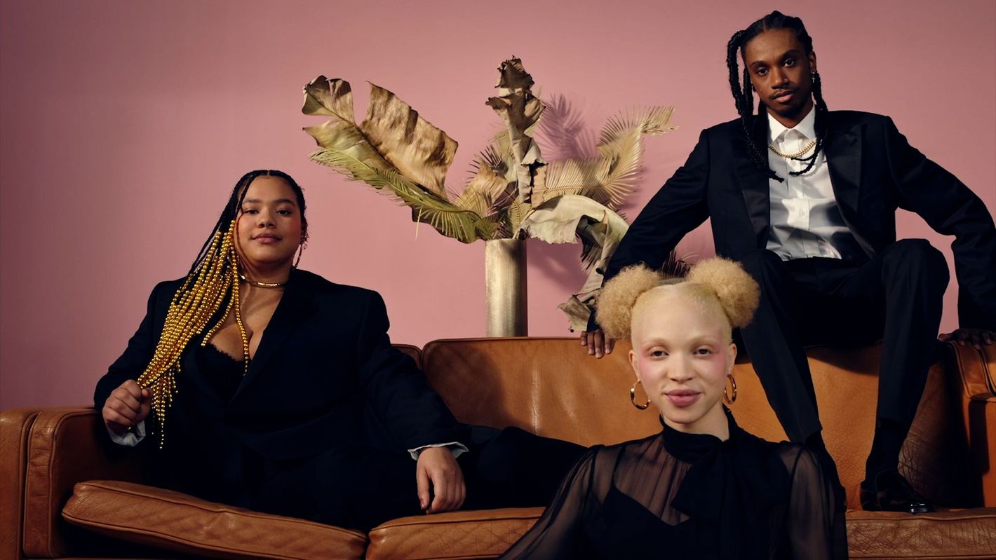 Three fashionably dressed people pose on a brown leather couch in front of a pale pink wall with a modern vase and dried palm leaves.