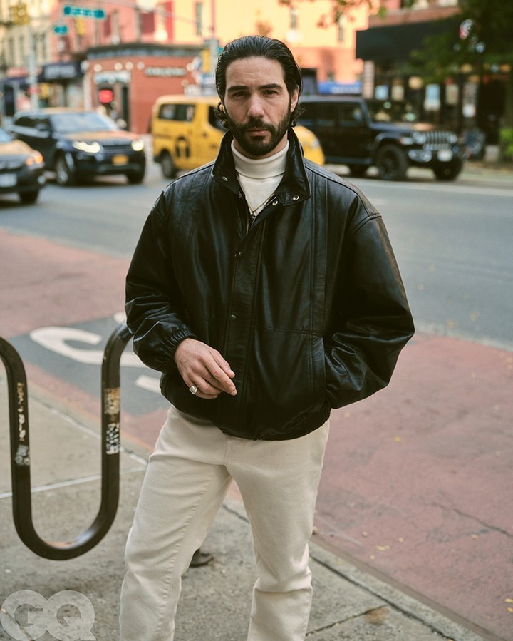 Adult male in black leather jacket and white turtleneck posing on a city street with buildings and traffic in the background.