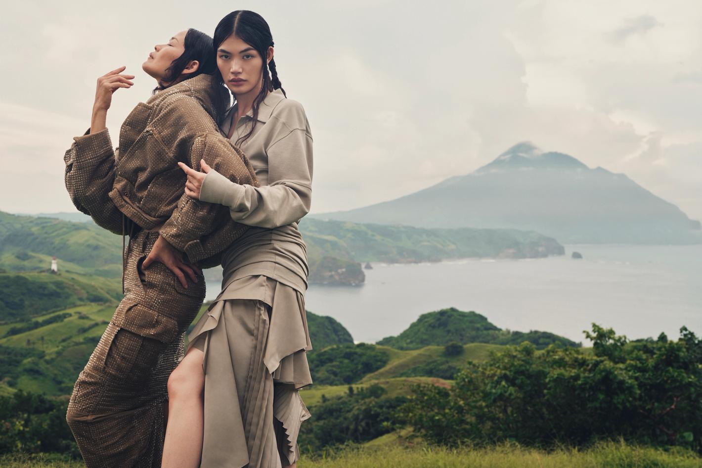 Two models in earth-toned apparel posing outdoors against a lush green rolling landscape and a mountain in the background.