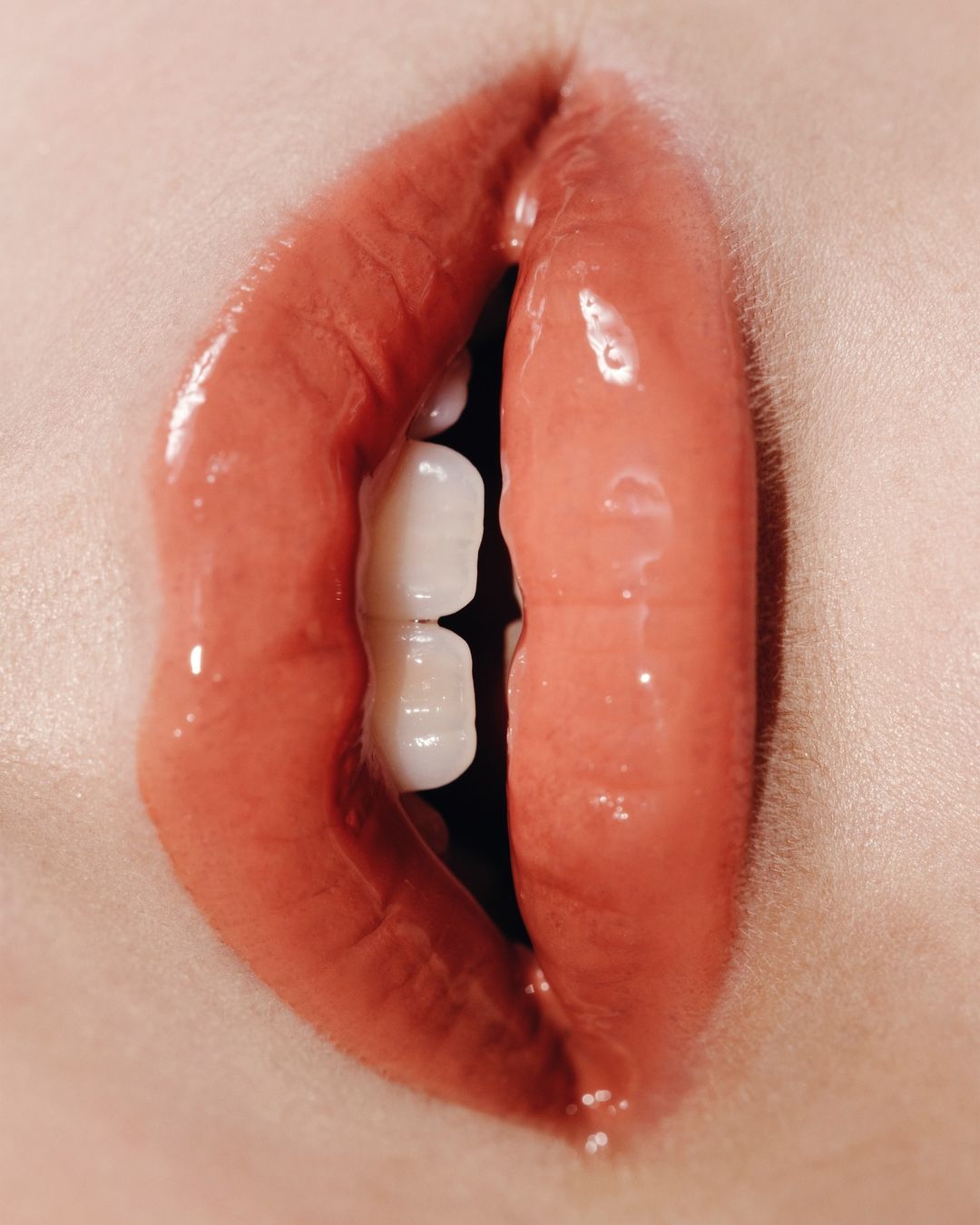Close-up studio shot of a person's open mouth featuring glossy coral-red lips, white teeth, and the surrounding skin.