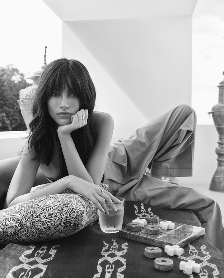 Black and white portrait of a young woman with bangs lounging on a sofa with a patterned pillow, side table, and a glass of water.