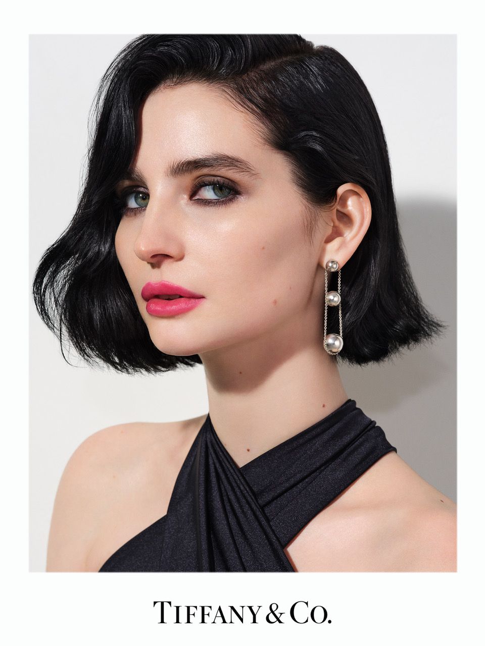 Elegant portrait of a brunette woman with pink lipstick wearing pearl earrings and a black dress, set against a clean white backdrop.