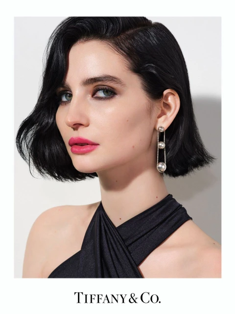 Elegant portrait of a brunette woman with pink lipstick wearing pearl earrings and a black dress, set against a clean white backdrop.