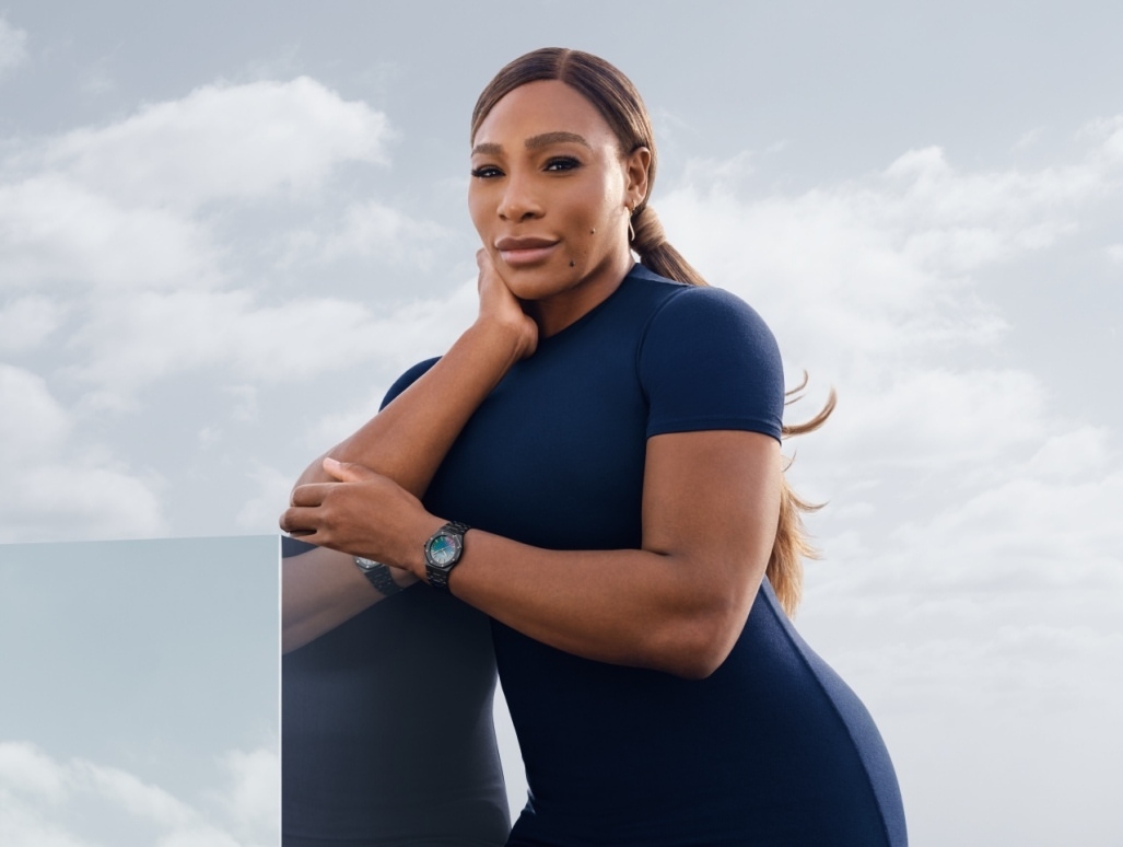 Portrait of Serena Williams in a navy dress posing outdoors against a cloudy sky with a sleek wristwatch and a reflective foreground surface.