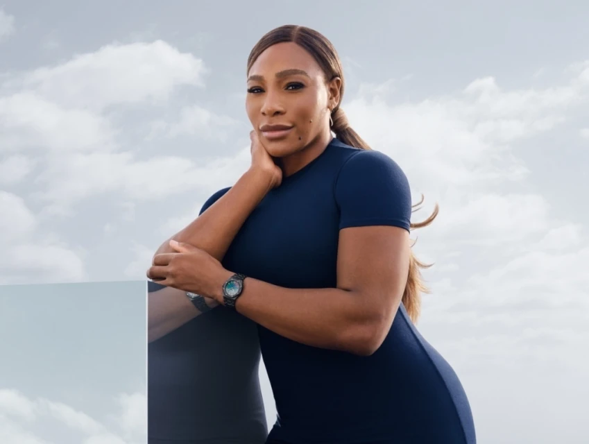 Portrait of Serena Williams in a navy dress posing outdoors against a cloudy sky with a sleek wristwatch and a reflective foreground surface.