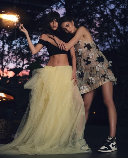 Two women pose together outdoors at dusk. One holds a sparkler, wearing a black top and yellow tulle skirt, and the other wears a dress and sneakers.