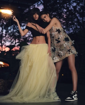 Two women pose together outdoors at dusk. One holds a sparkler, wearing a black top and yellow tulle skirt, and the other wears a dress and sneakers.