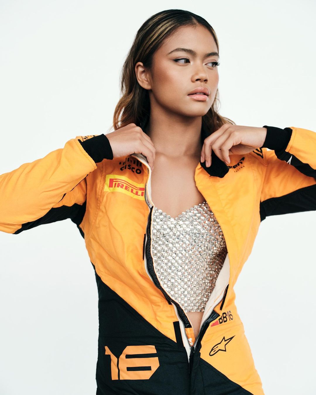 Woman poses in a yellow and black racing suit with a sparkling silver top in a studio shot against a bright white backdrop.