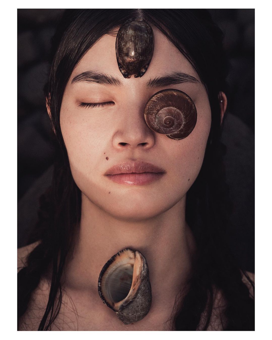 Artistic portrait of a woman with one eye closed, adorned with seashells on her face and neck, set against a blurred dark background.