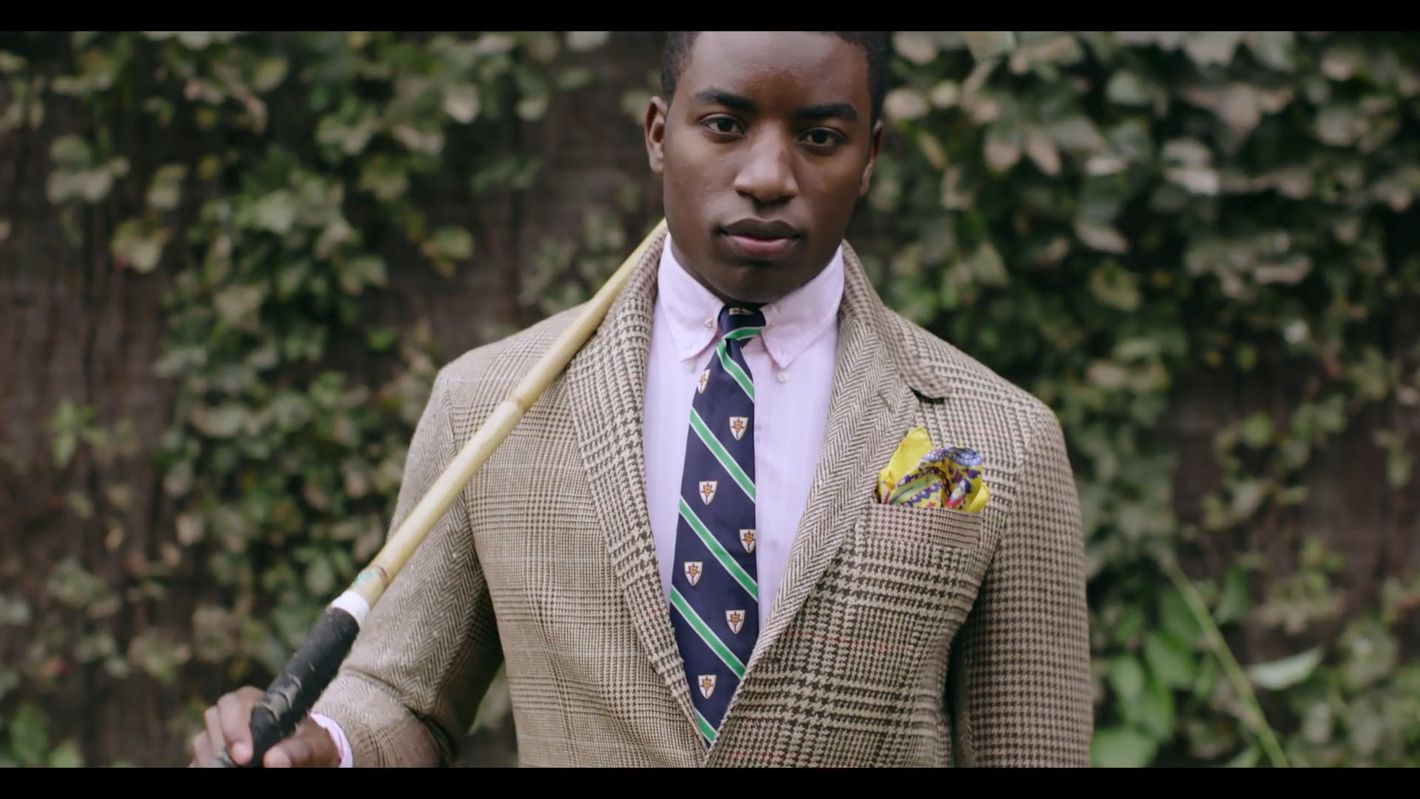Handsome man in blazer and tie holds a polo stick outdoors amid green foliage, projecting classic style and athletic elegance.