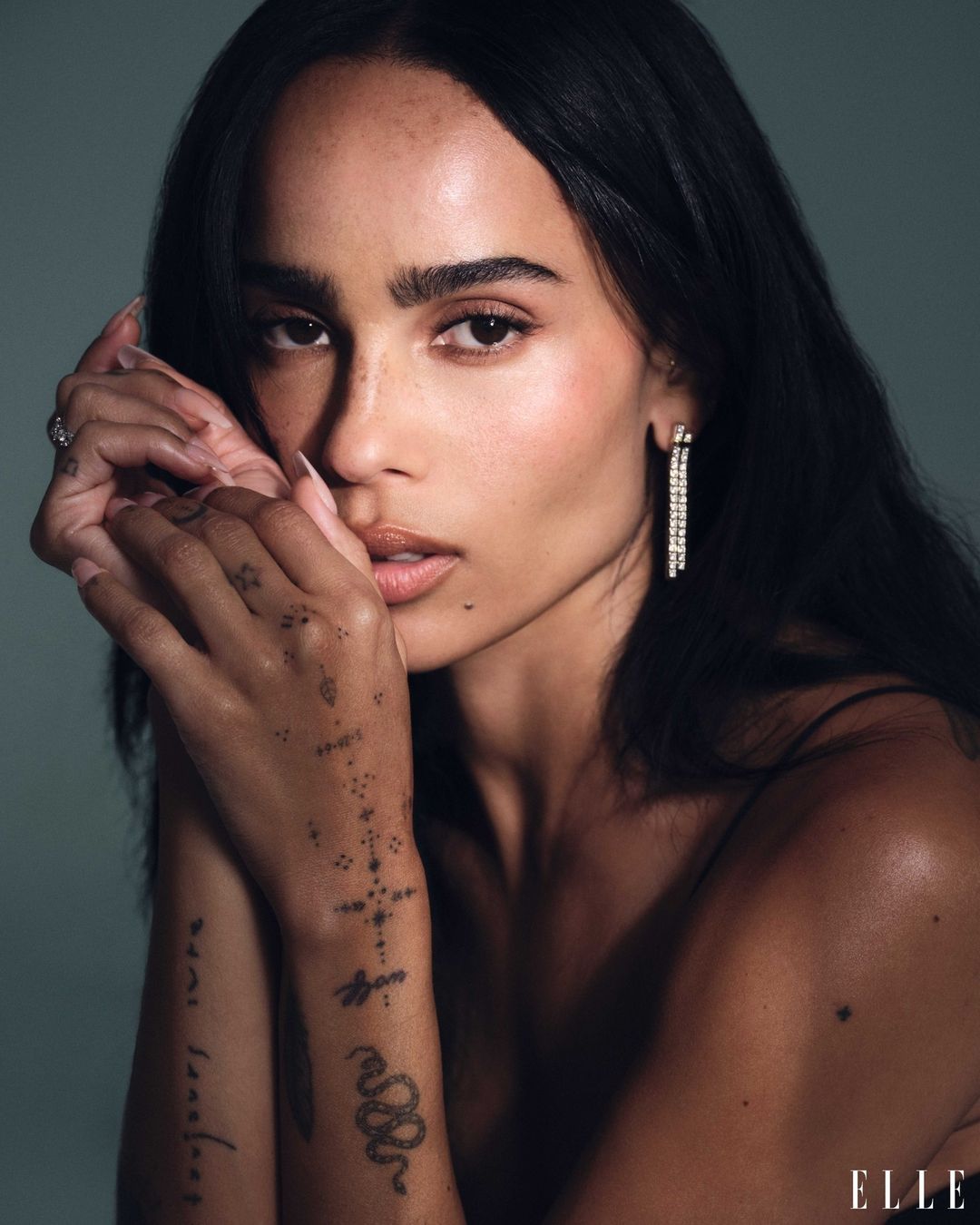 Close-up portrait of Zoë Kravitz with dark hair, tattoos on her hands, and drop earrings against a neutral background.