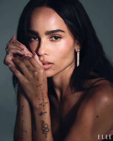 Close-up portrait of Zoë Kravitz with dark hair, tattoos on her hands, and drop earrings against a neutral background.