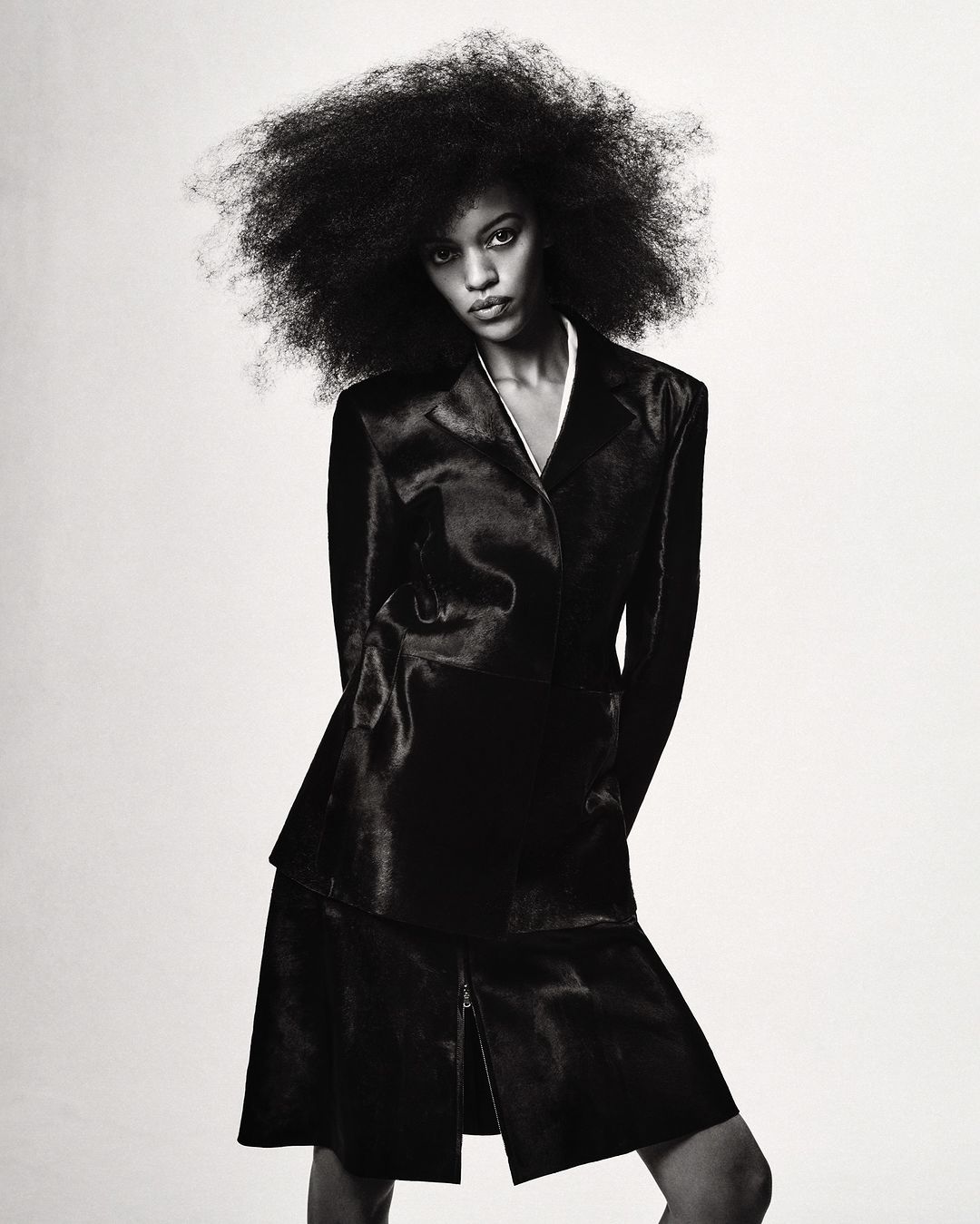 Model with voluminous dark curly hair wearing a black jacket and matching skirt against a plain background.