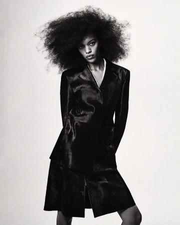 Model with voluminous dark curly hair wearing a black jacket and matching skirt against a plain background.