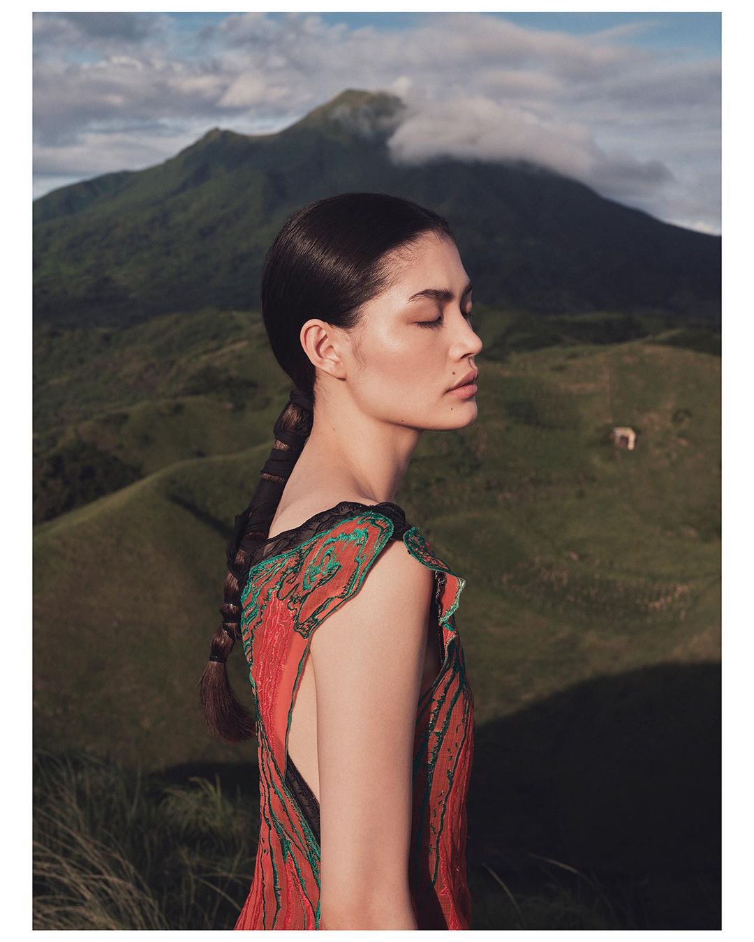 Elegant woman posing outdoors against mountain backdrop, wearing sleeveless coral dress with braided hair, eyes closed, serene and natural.