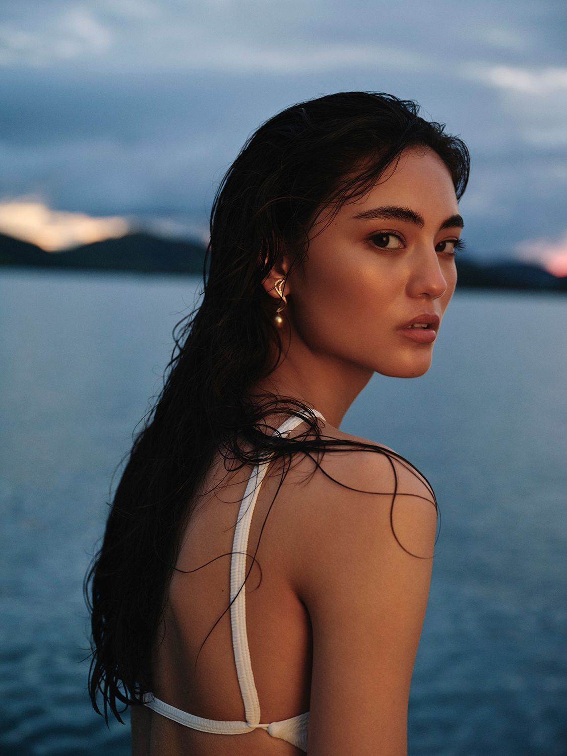 Portrait of a young woman with dark hair and pearl earrings looking over her shoulder with the ocean and sunset sky in the background.
