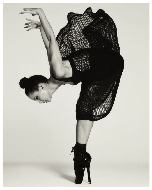 A ballet dancer balances en pointe in a black dress. The dancer strikes a dramatic pose against a light grey background.
