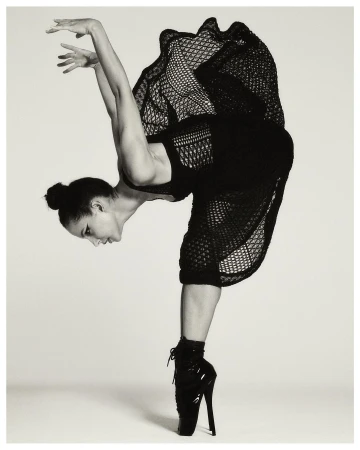 A ballet dancer balances en pointe in a black dress. The dancer strikes a dramatic pose against a light grey background.