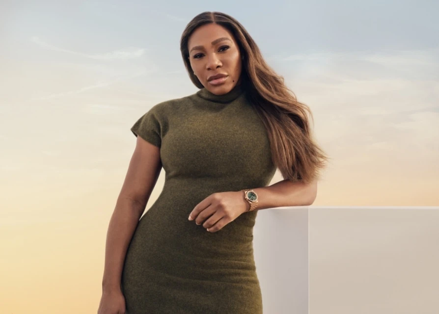 Serena Williams poses in olive green dress with a gold wristwatch against a gradient sky backdrop, leaning on a white surface.