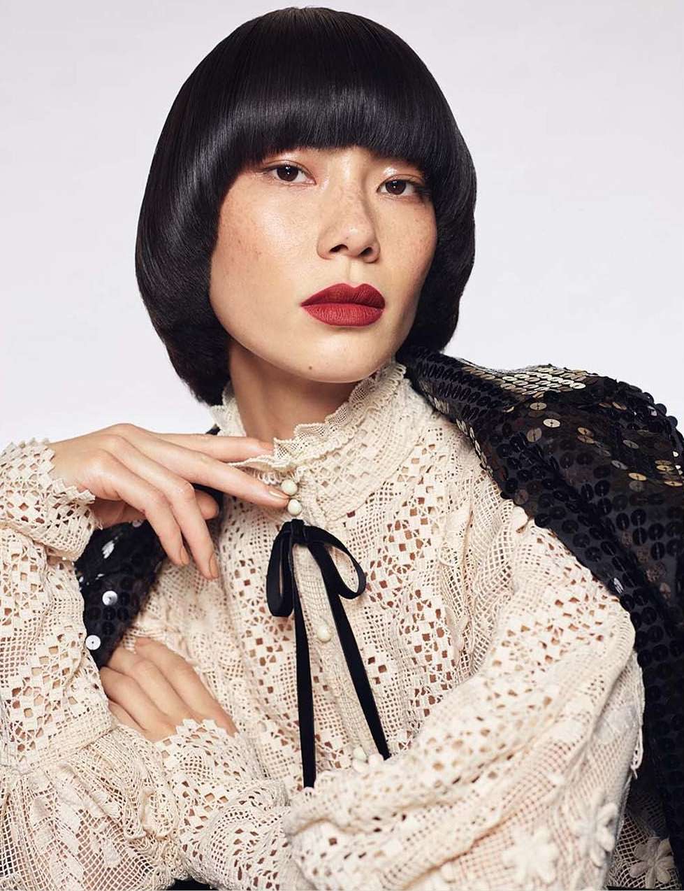 Asian woman in eyelet blouse and sequined jacket poses in a studio with short, dark hair and dark red lipstick. Beauty and fashion.
