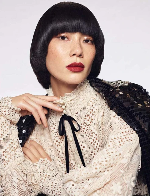 Asian woman in eyelet blouse and sequined jacket poses in a studio with short, dark hair and dark red lipstick. Beauty and fashion.