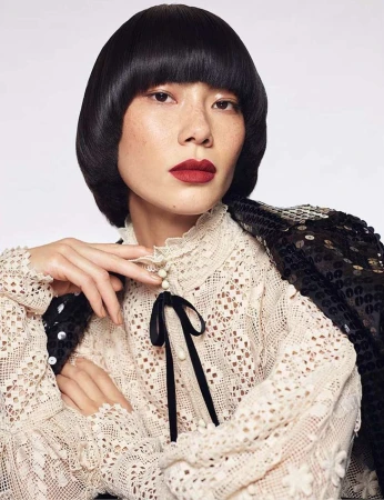 Asian woman in eyelet blouse and sequined jacket poses in a studio with short, dark hair and dark red lipstick. Beauty and fashion.