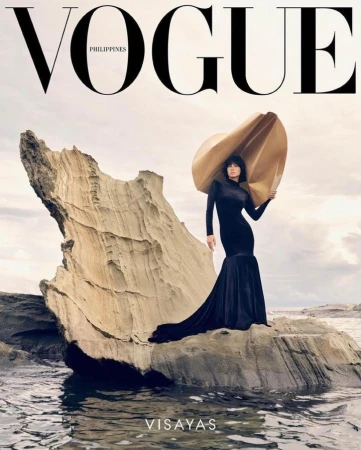 A woman in a long black dress and large sculptural hat standing on a rocky outcrop surrounded by water.