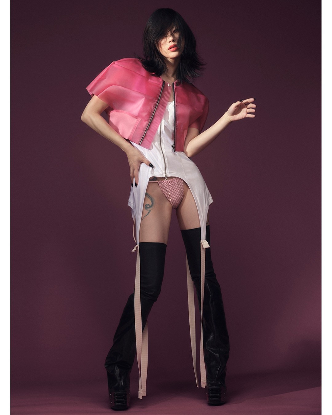 Fashion photo of a slender adult female in a translucent jacket, white dress, and thigh-high boots striking a pose on a plum background.