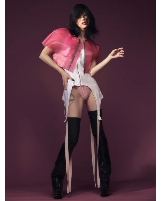 Fashion photo of a slender adult female in a translucent jacket, white dress, and thigh-high boots striking a pose on a plum background.