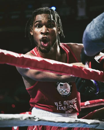 African-American male boxer in red Olympic team uniform stands in a boxing ring with an excited expression, mouth open, and focused gaze.