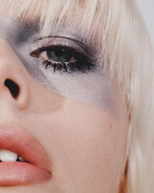 Close-up portrait of a woman with platinum blonde hair, silver eyeshadow, and pink lips against a solid white background.