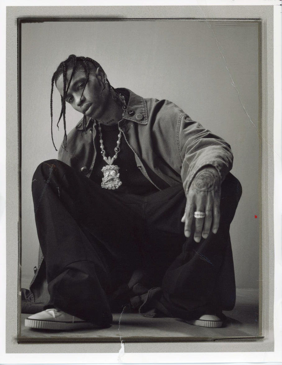 Monochromatic portrait of adult male in jacket and dark pants crouching indoors with visible jewelry, tattoos, and casual shoes.