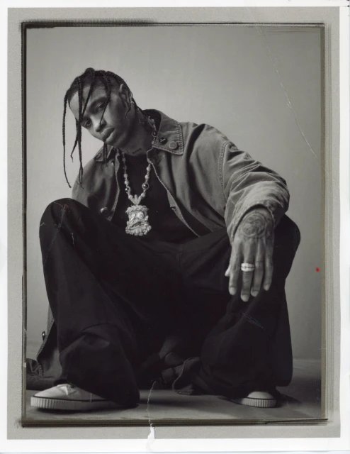 Monochromatic portrait of adult male in jacket and dark pants crouching indoors with visible jewelry, tattoos, and casual shoes.