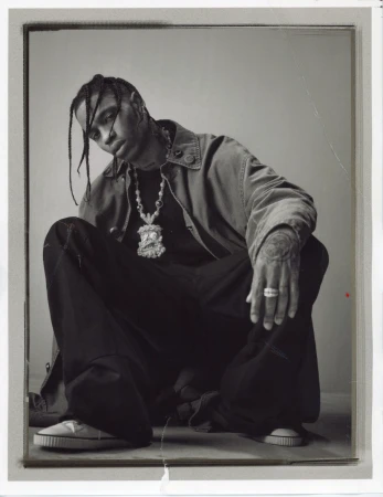 Monochromatic portrait of adult male in jacket and dark pants crouching indoors with visible jewelry, tattoos, and casual shoes.