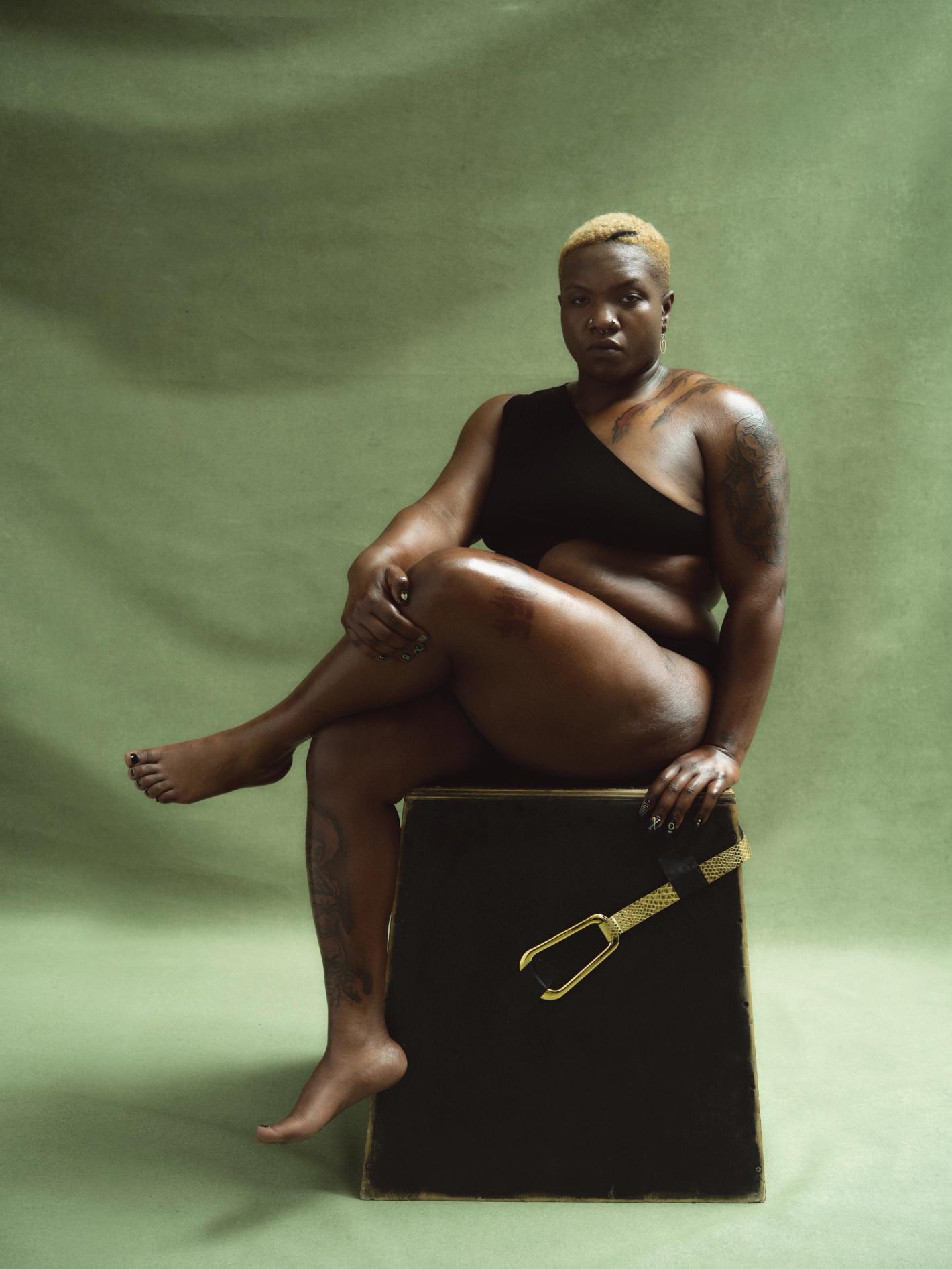 Portrait of a confident Black woman with short blonde hair sitting on a black cube with a gold belt against a green backdrop.