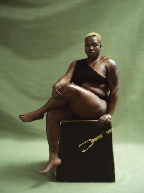 Portrait of a confident Black woman with short blonde hair sitting on a black cube with a gold belt against a green backdrop.