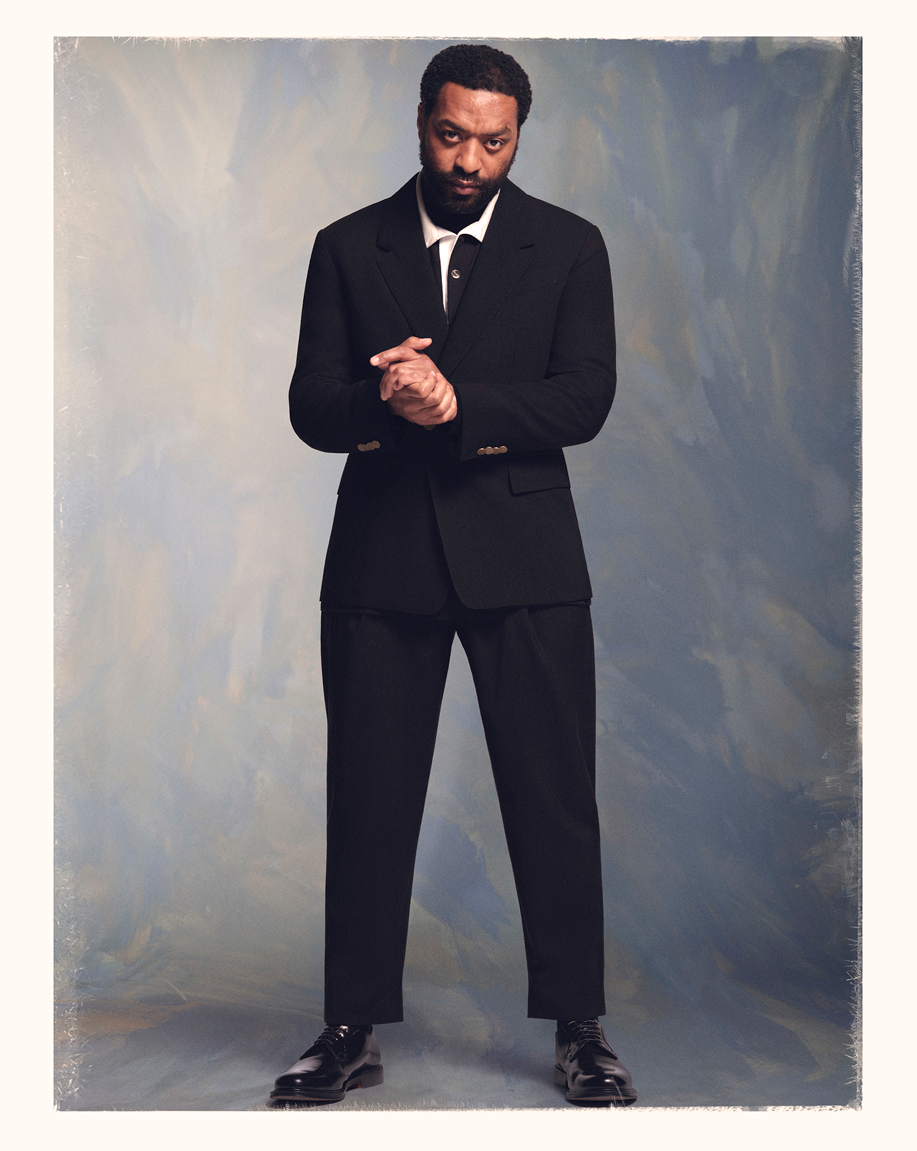 Full-length portrait of a Black man in a black suit with his hands clasped against a textured painted backdrop
