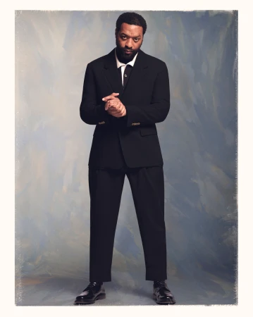 Full-length portrait of a Black man in a black suit with his hands clasped against a textured painted backdrop