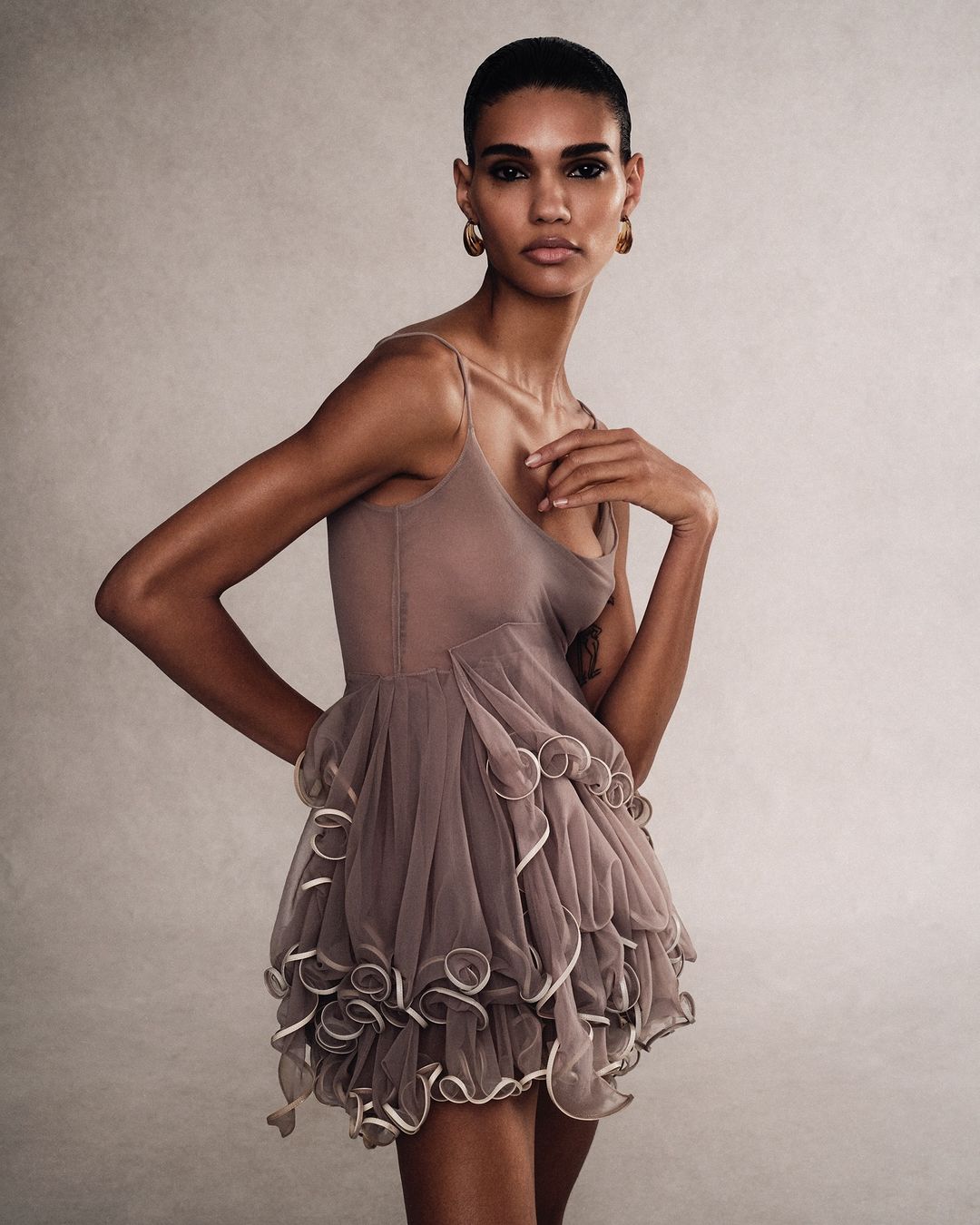 Elegant fashion portrait of a woman with short dark hair posing in a stylish light brown ruffled dress with spaghetti straps in a studio.