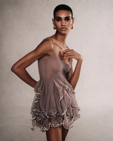 A woman wearing a taupe slip dress with a ruffled skirt standing against a textured grey studio backdrop.