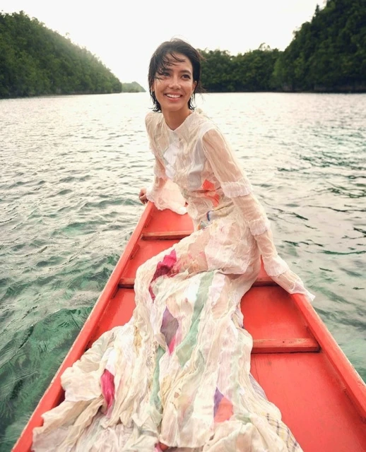 Smiling woman in long dress sits in a red wooden boat on shimmering water with green, tree-covered isles in the background.