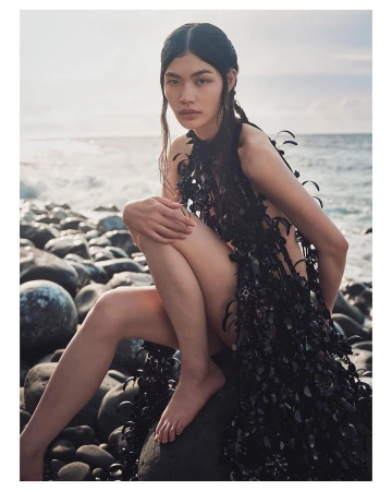Asian woman in dark textured sleeveless dress perched on dark round boulders near an ocean shore by Sharif Hamza