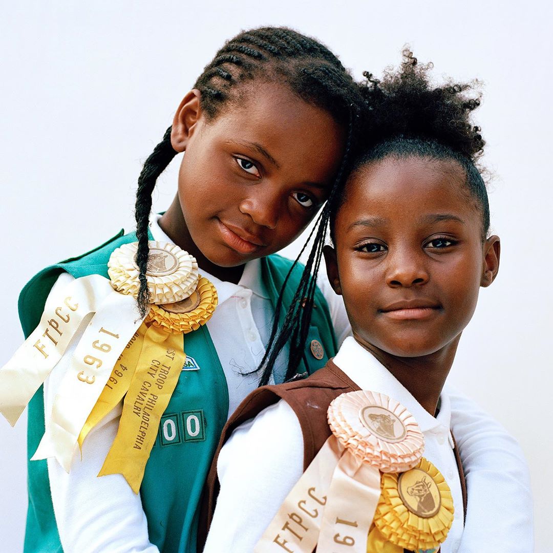 Portrait of two young African-American girls in scouting uniforms with award ribbons posing closely against a bright, clean background.
