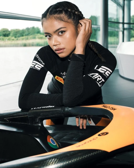 Woman with braids posing in a black top leaning against a race car with sponsor logos, in an indoor setting with natural lighting.