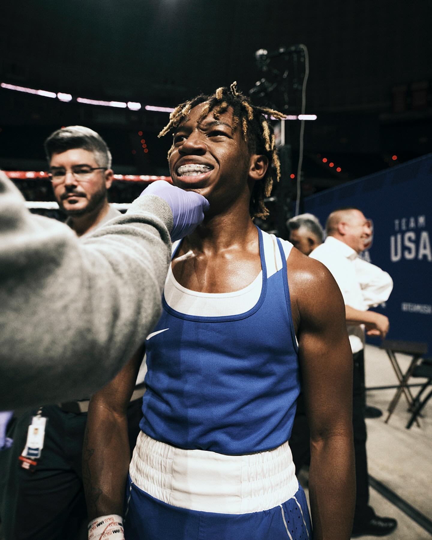 Close-up of an African-American boxer with braces being examined by a medic, with two other men visible in the background inside.