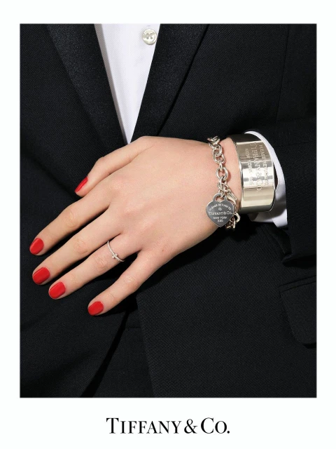 Close-up of a person's hand and wrist adorned with a silver charm bracelet and a thin silver ring, set against a dark suit.