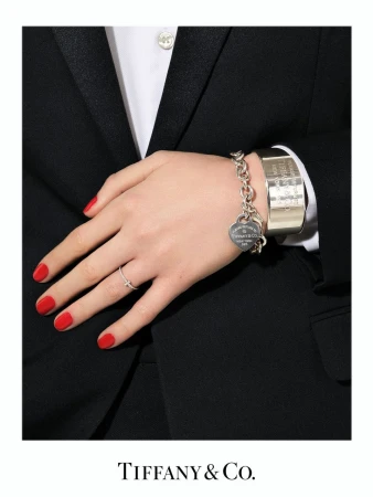 Close-up of a person's hand and wrist adorned with a silver charm bracelet and a thin silver ring, set against a dark suit.