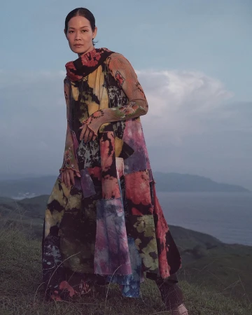 Woman wearing a long patterned patchwork dress standing on a grassy coastal hill overlooking the sea